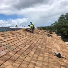 Workers installing shingles on a large roof under a cloudy sky.